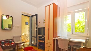 Ein kompakter Wohnraum mit Küchenzeile, Essbereich, Fernseher und einem großen Fenster mit Blick ins Grüne.