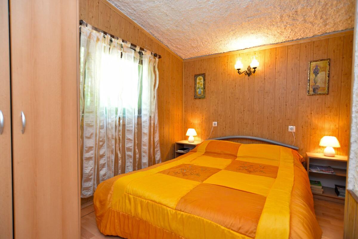 A room with a bed, wooden walls, a window with curtains, and two wall-mounted lamps.