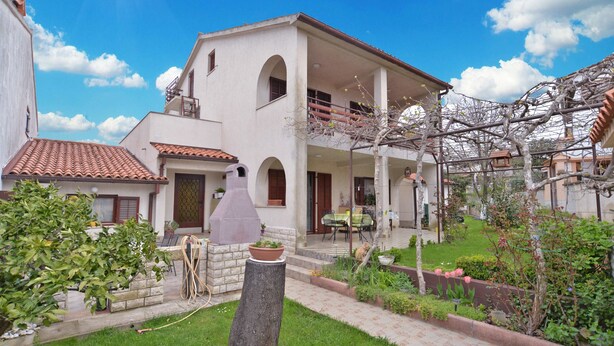 Une maison à deux étages avec un balcon, un jardin agrémenté d’un tronc d’arbre et une pergola couverte de plantes grimpantes.