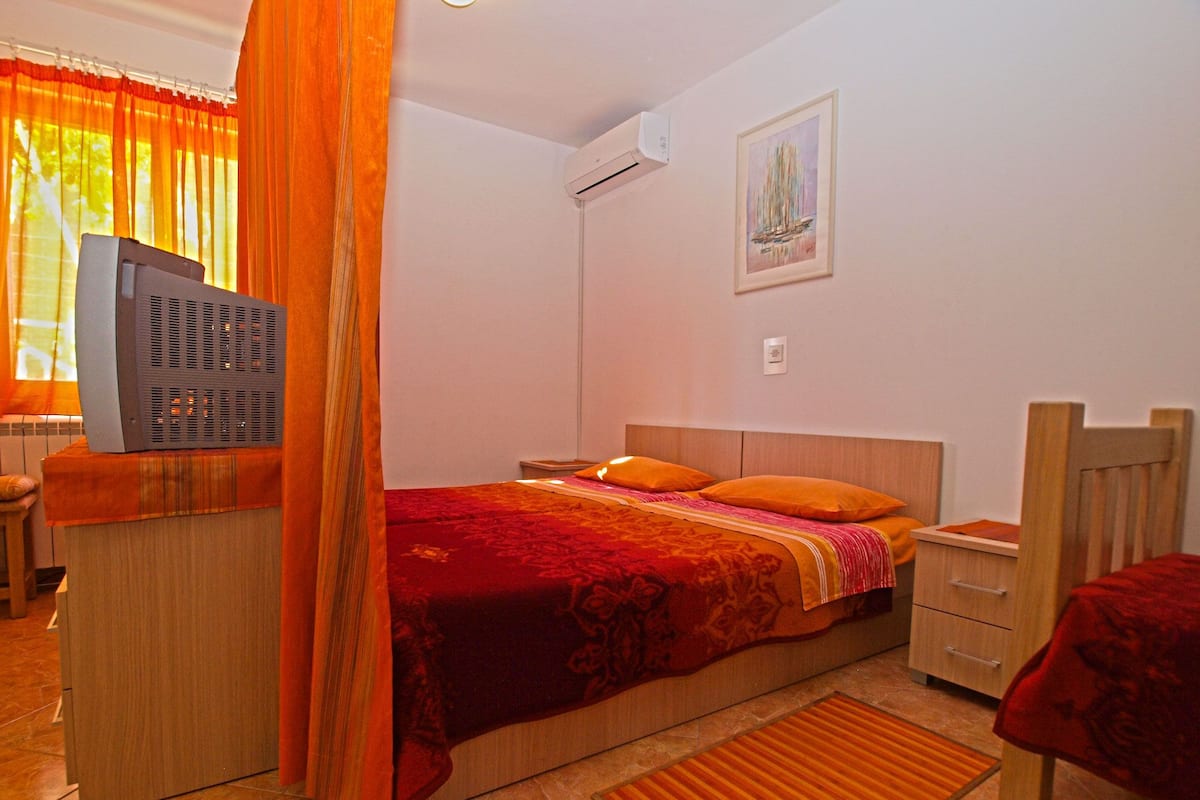 Quarto com cama, mesa de cabeceira, televisão, janela com cortinas, aparelho de ar condicionado e um quadro na parede.