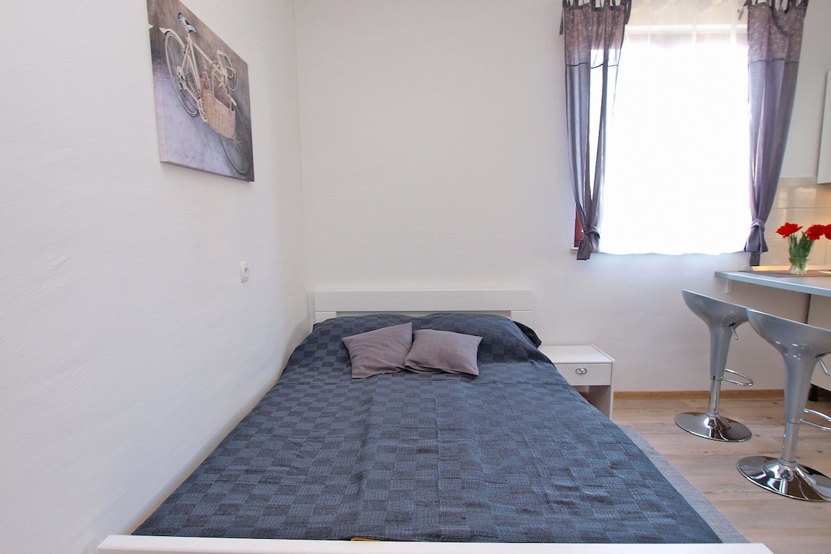 Quarto pequeno com uma cama de solteiro, cabeceira branca, mesa de cabeceira e uma cadeira.