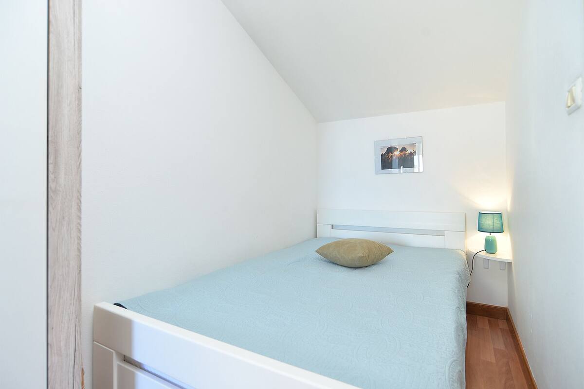 Quarto pequeno com uma cama de solteiro, teto inclinado, mesa de cabeceira com abajur e um quadro emoldurado na parede.