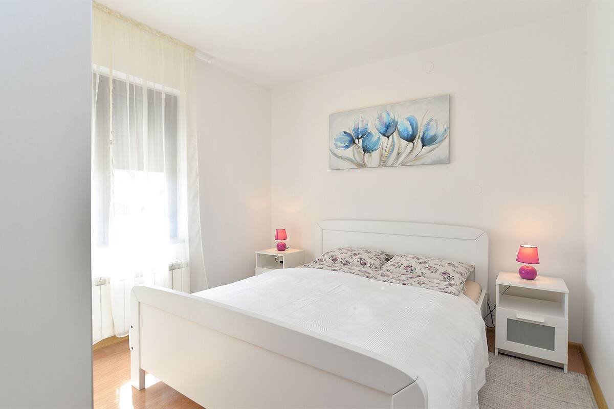 Quarto com cama branca, duas mesas de cabeceira com luminárias, janela com cortinas leves e um quadro floral na parede.