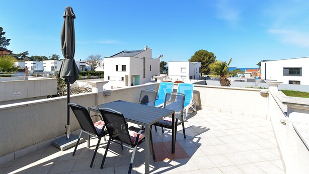 A terrace with a table and chairs, a sun umbrella, and a view of modern buildings and the sea. - Apartment 589 (Fazana)