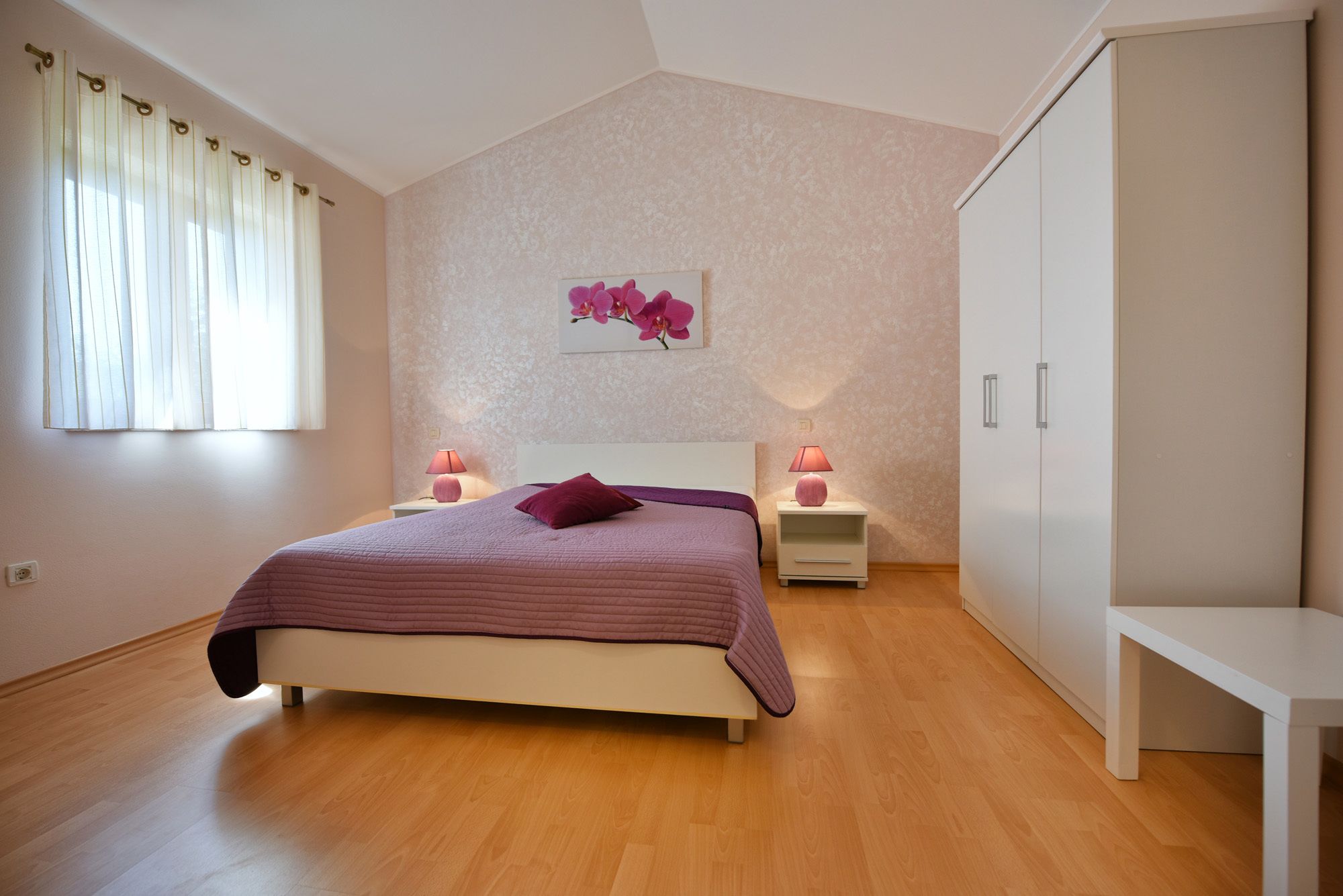 Ein Schlafzimmer mit einem Bett, Nachttischen mit Lampen, einem Kleiderschrank und einem Fenster mit Vorhängen.