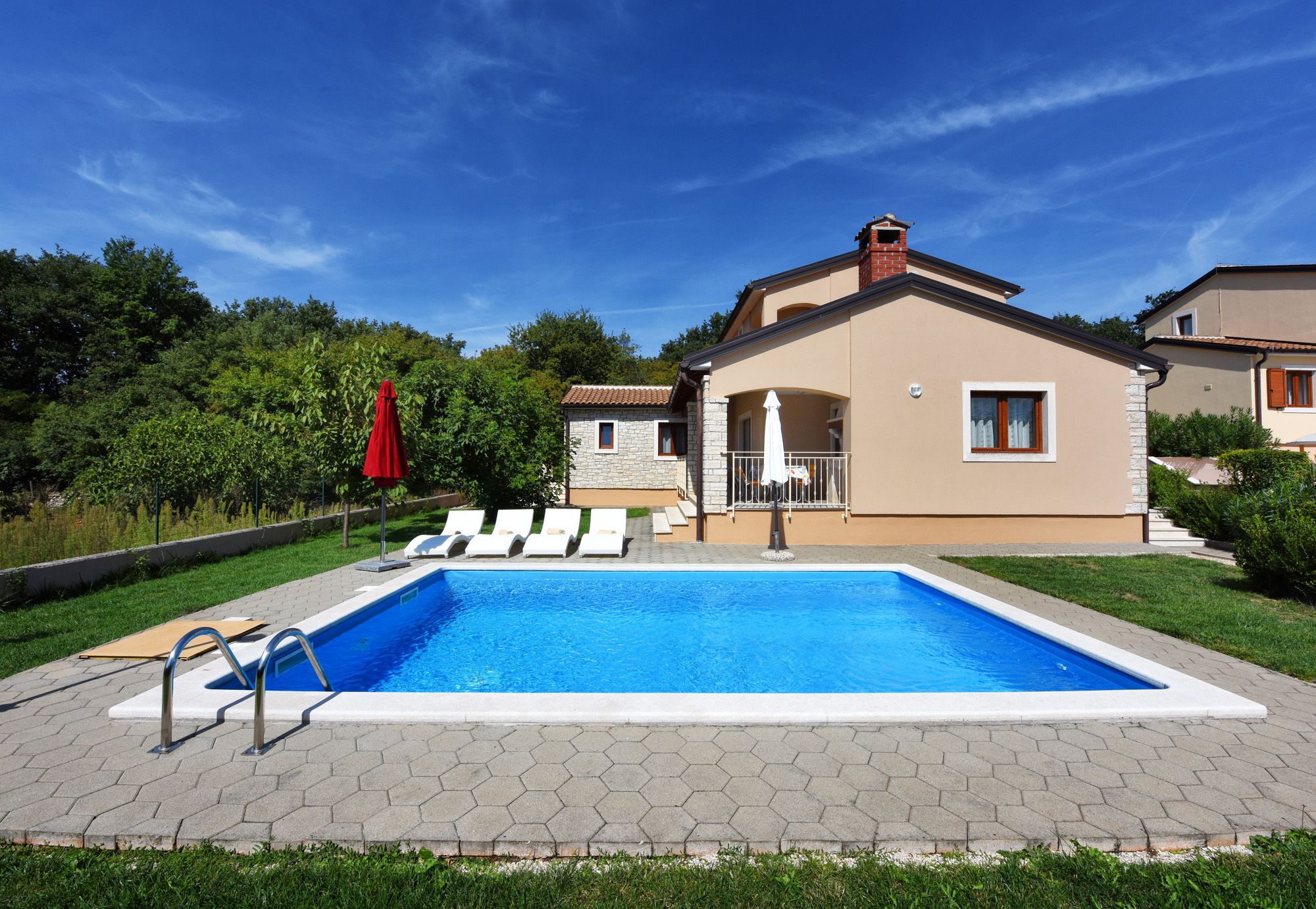 A swimming pool with a tiled deck, a red umbrella, and lounge chairs.