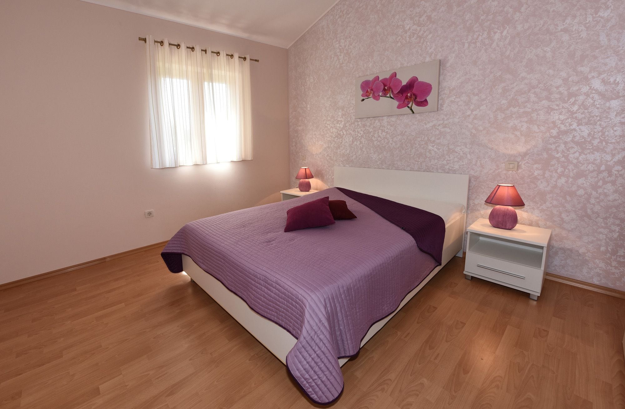 Une chambre à coucher avec un lit, deux tables de chevet, une fenêtre avec des rideaux et un tableau représentant une orchidée accroché au mur.