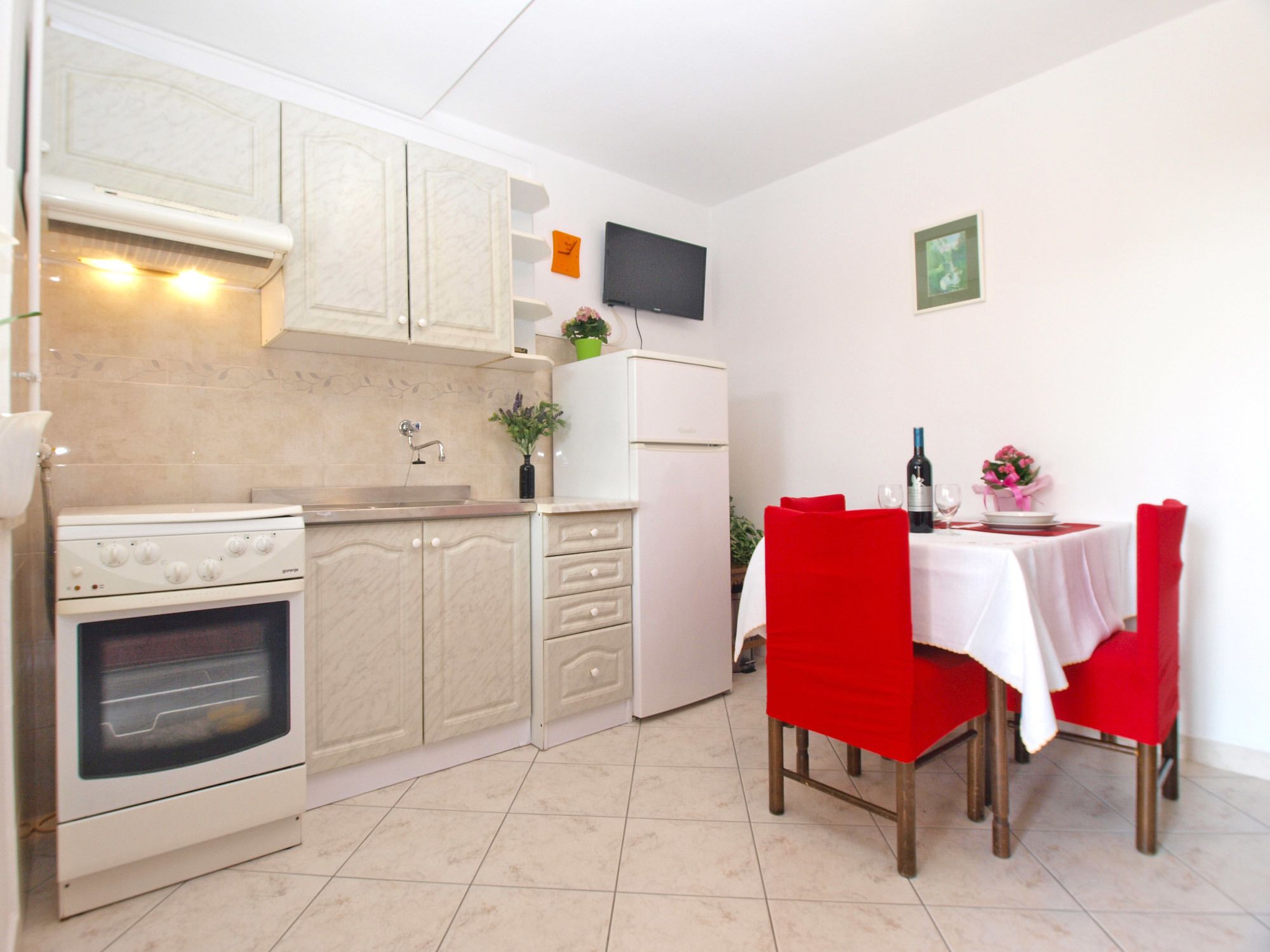 A compact kitchen with a stove, oven, and white cabinets. A dining table with red chairs and a white tablecloth is set up for a meal.