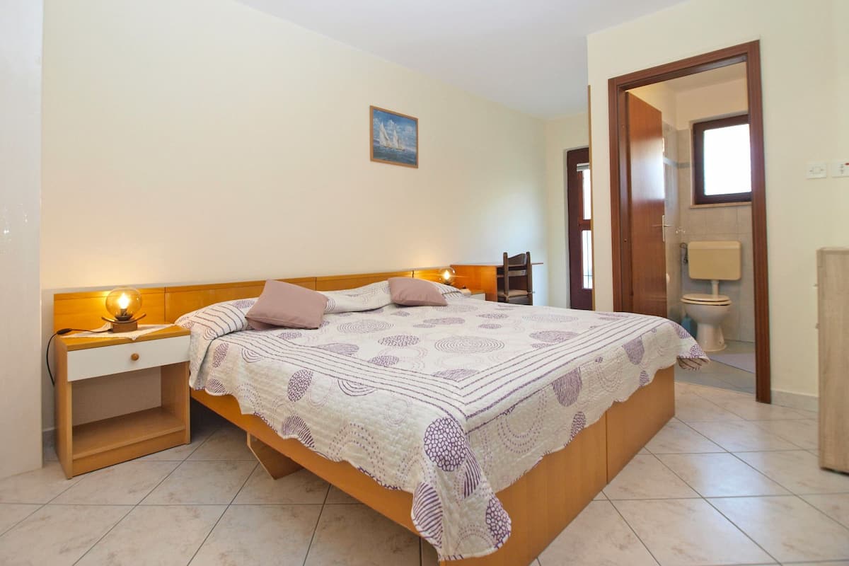 A bedroom with a bed, bedside tables, a nightstand, a chair, a painting on the wall, and a door leading to a bathroom.