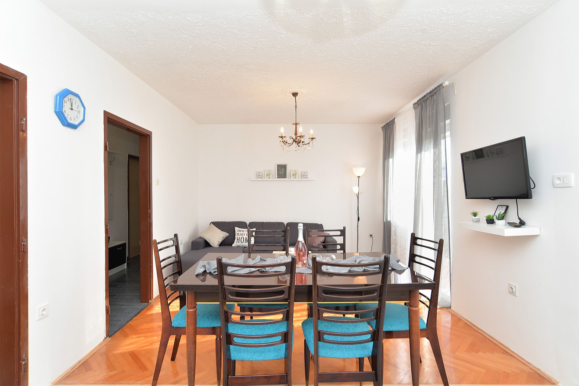 A dining area with a table and chairs, a chandelier, a television on the wall, and a sofa in the background.