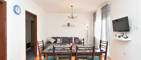A dining area with a table and chairs, a chandelier, a television on the wall, and a sofa in the background.