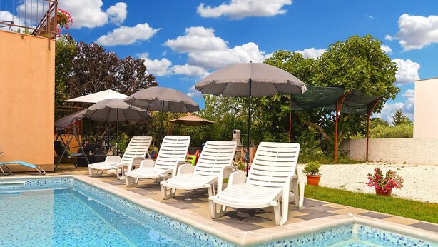 Zona de piscina con sillones de descanso y sombrillas, cielo azul despejado y follaje al fondo.