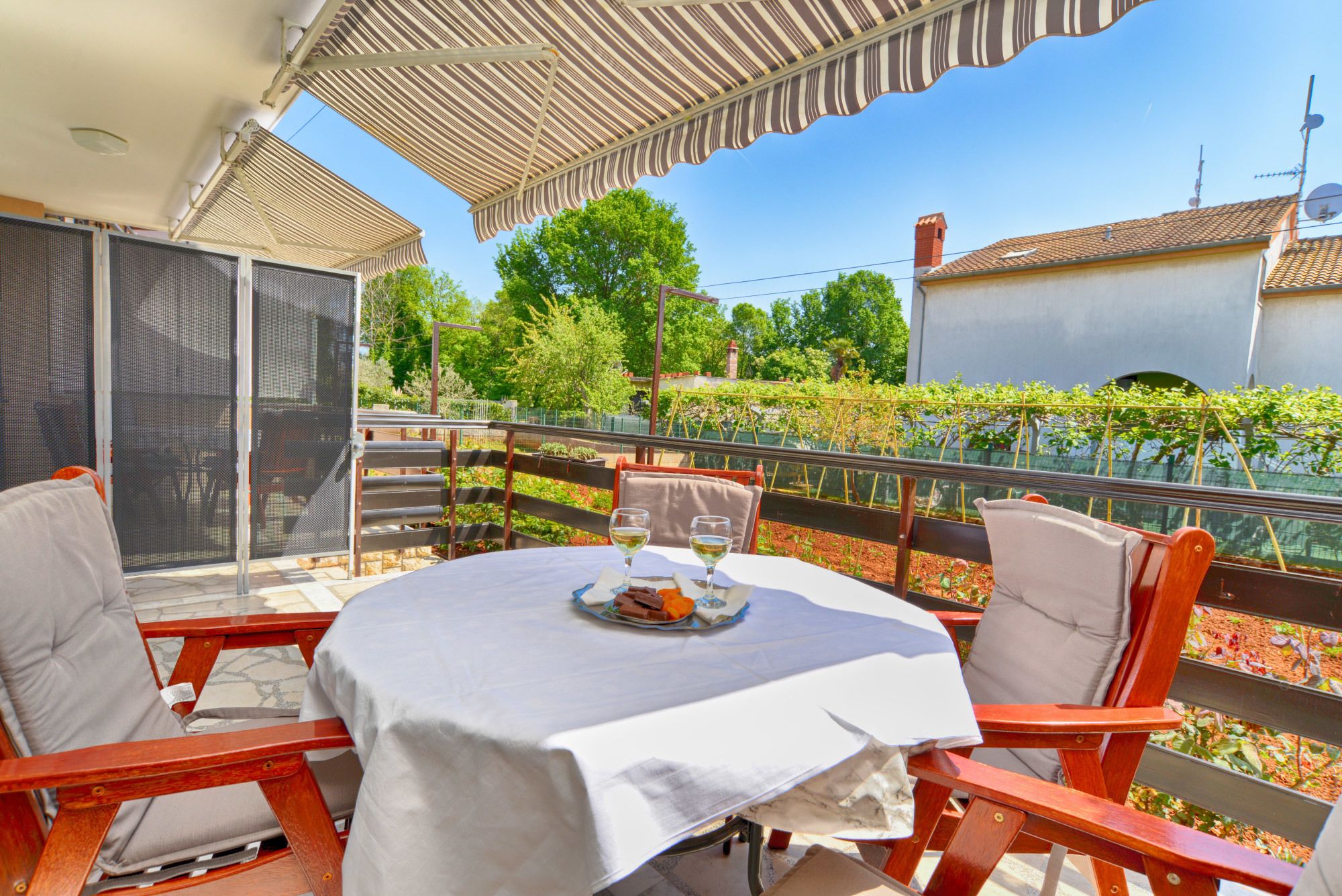 Patio esterno con tavolo apparecchiato per due, circondato da sedie di legno, con vista su un giardino e un vigneto.