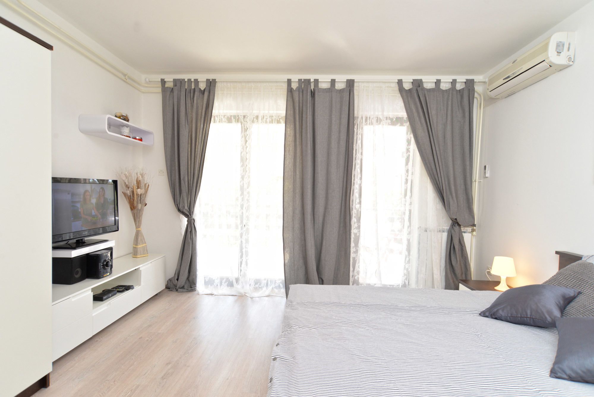 Ein Schlafzimmer mit einem Bett, einem Fernseher auf einem Schrank, einem Nachttisch mit Lampe und einem Fenster mit Vorhängen.