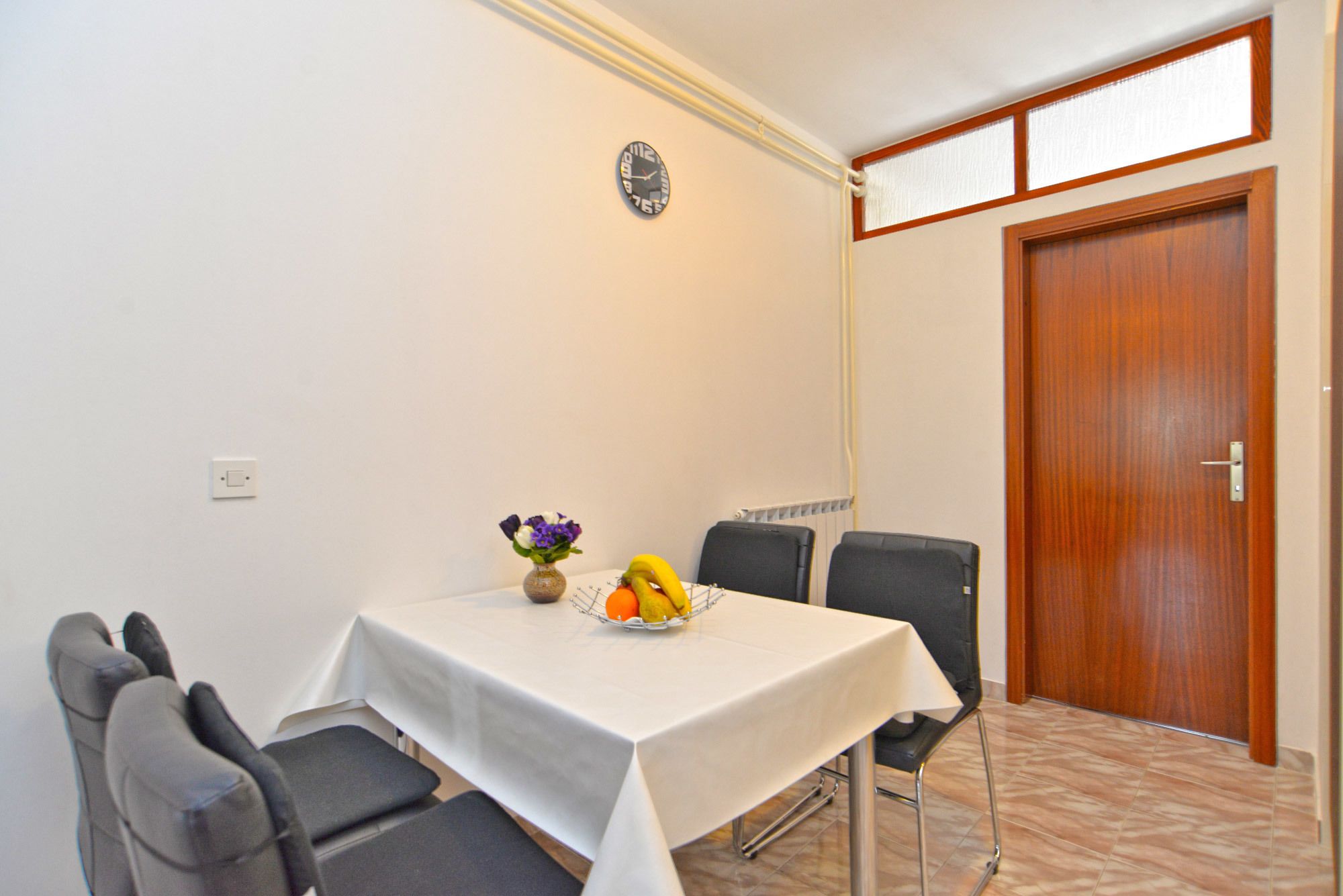 A dining area with a table set for four, a clock on the wall, and a wooden door.