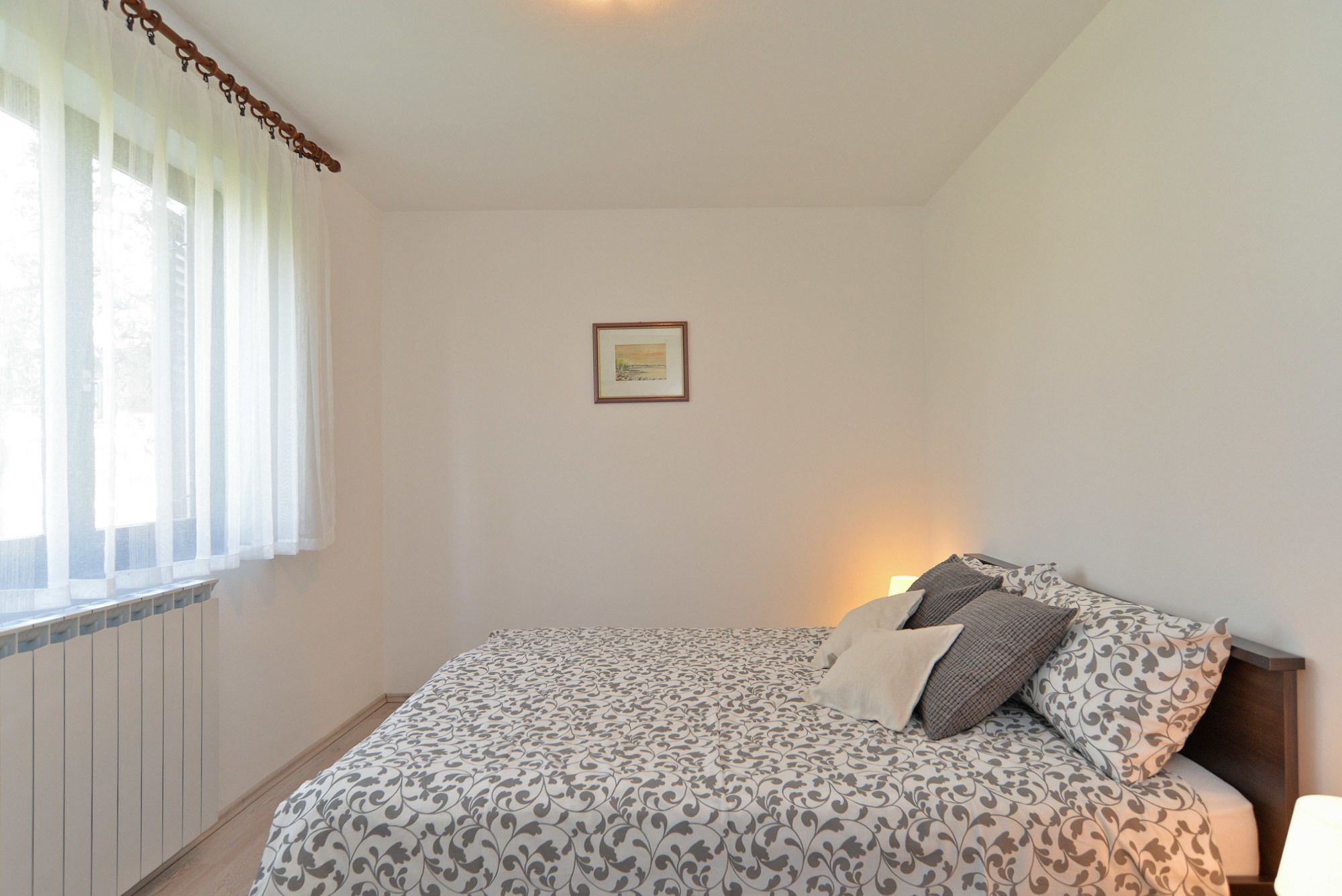 A bedroom with a bed, a bedside lamp, a framed picture on the wall, and a window with sheer curtains.