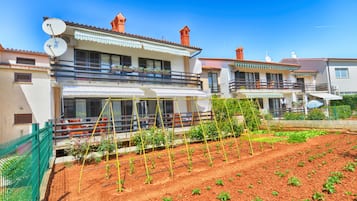 A residential building with a balcony, a garden with young plants, and a clear blue sky.