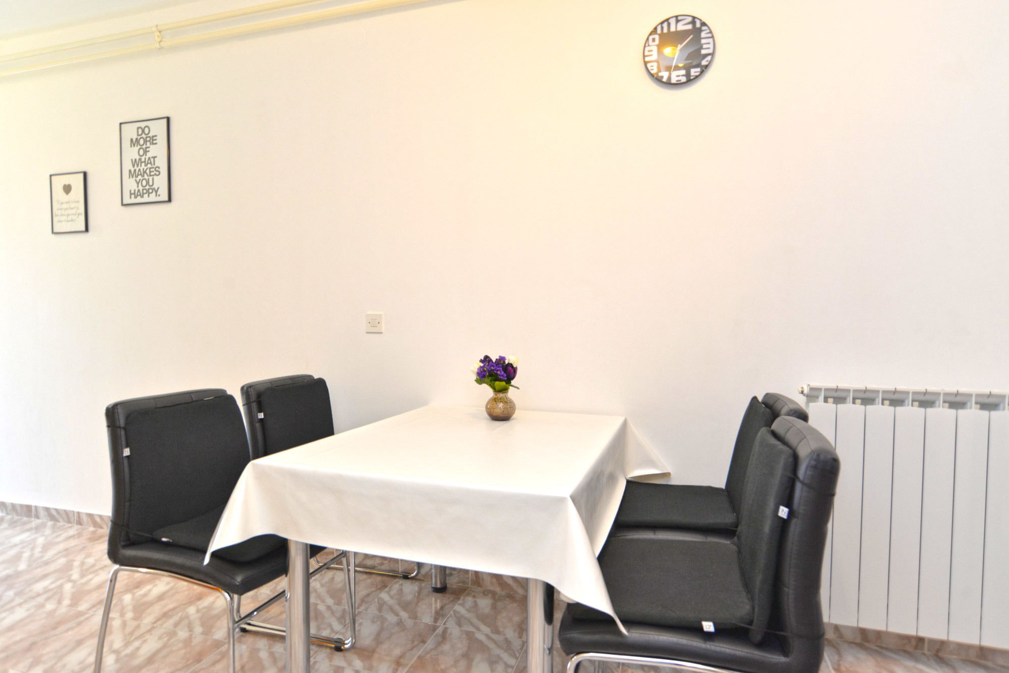 Une salle de conférence avec une table blanche, quatre chaises noires, un petit vase avec des fleurs violettes et une horloge au mur.