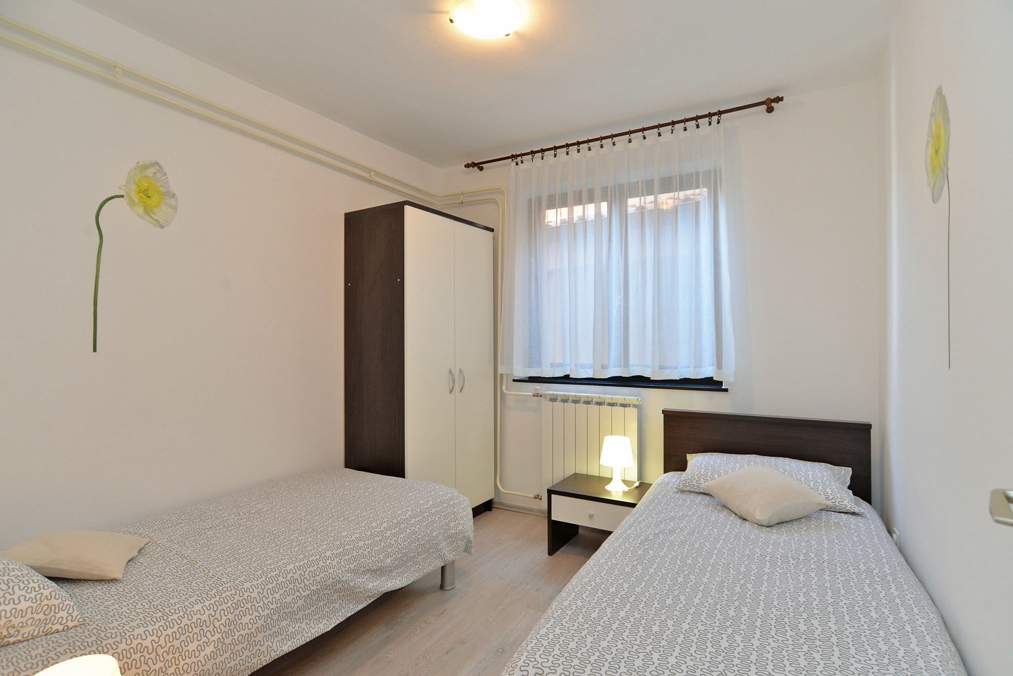 Ein Schlafzimmer mit zwei Einzelbetten, einem Nachttisch mit Lampe, einem Kleiderschrank und einem Fenster mit transparenten Vorhängen.