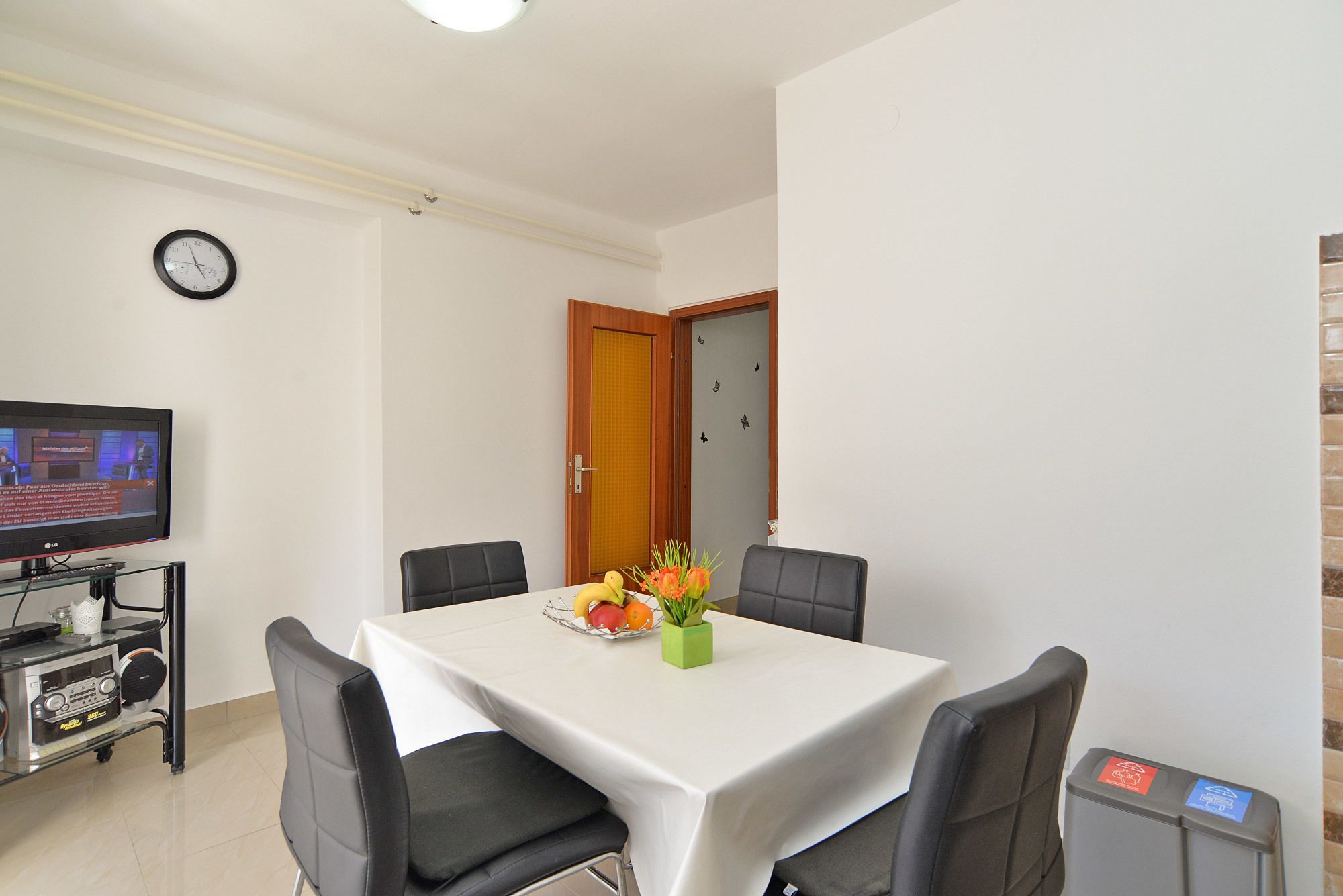 Un comedor con una mesa blanca, cuatro sillas, un televisor sobre un mueble bajo y una planta pequeña sobre la mesa.