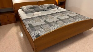 A wooden bed with a patterned bedspread and pillows, a bedside table, and a wall-mounted shelf. - Apartment 650 (Labin)
