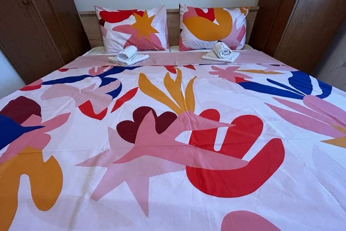 Una cama con una colcha abstracta y colorida, dos almohadas con diseños similares, y una cama bien hecha con una almohada blanca y una toalla blanca doblada.