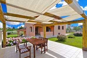 A covered outdoor patio with a wooden table and chairs, a tiled floor, and a view of a building and garden.