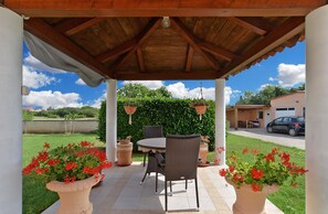A covered patio with a table and chairs, surrounded by potted plants and a view of a lake and buildings.
