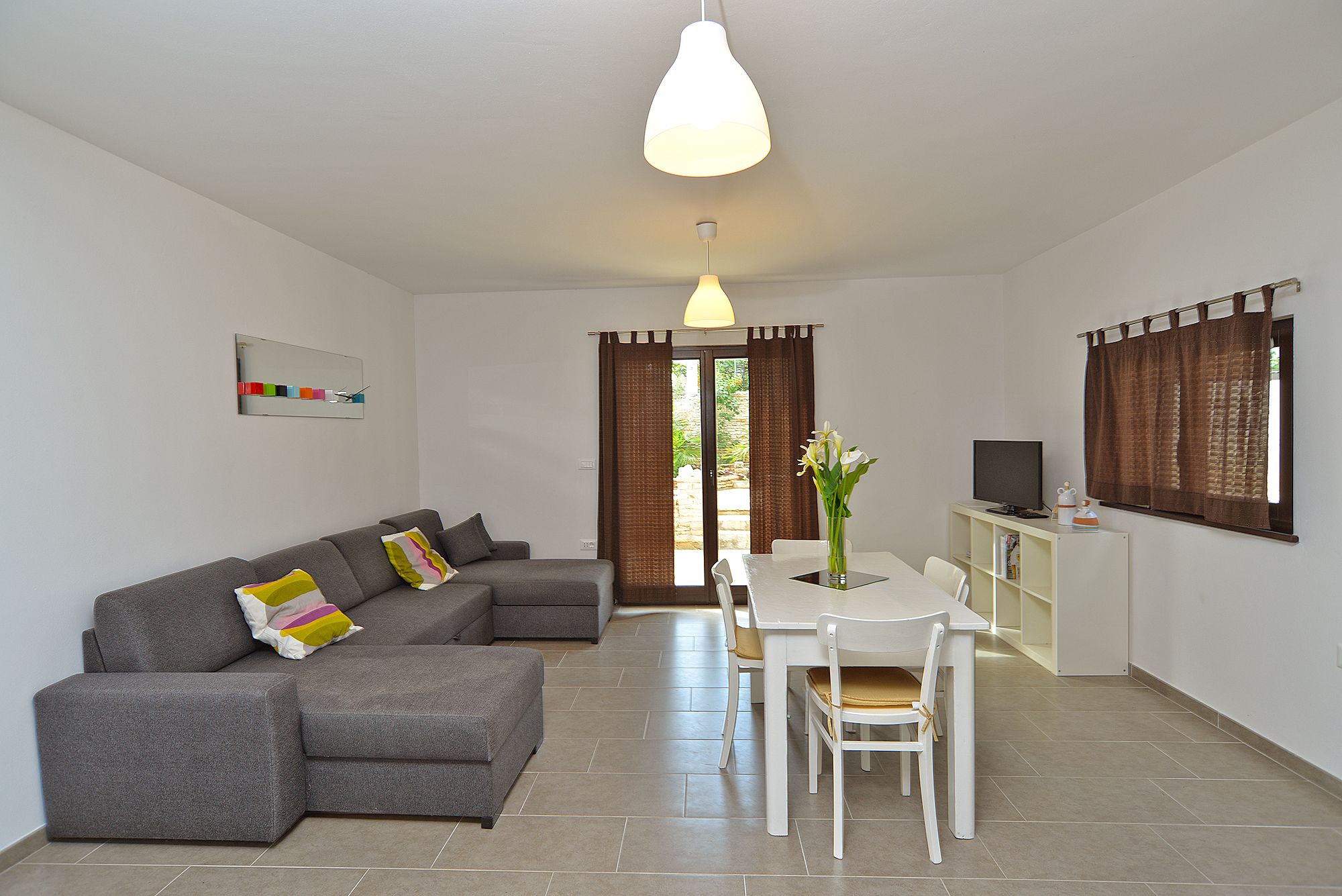 A modern living room with a grey sectional sofa, a white dining table with chairs, a television on a white cabinet, and a large window with brown curtains.