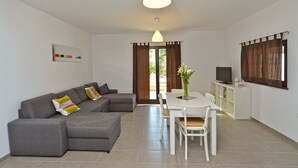 A modern living room with a grey sectional sofa, a white dining table with chairs, a television on a white cabinet, and a large window with brown curtains. - House 682 (Svetvincenat)