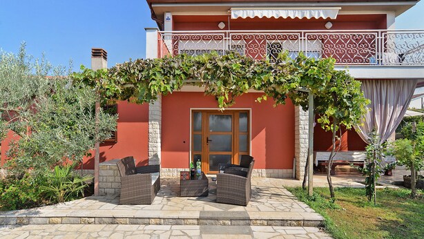Ein zweistöckiges Gebäude mit roter Fassade, einem Balkon mit weißen Geländern und einer überdachten Terrasse mit Korbmöbeln und einem kleinen Tisch.
