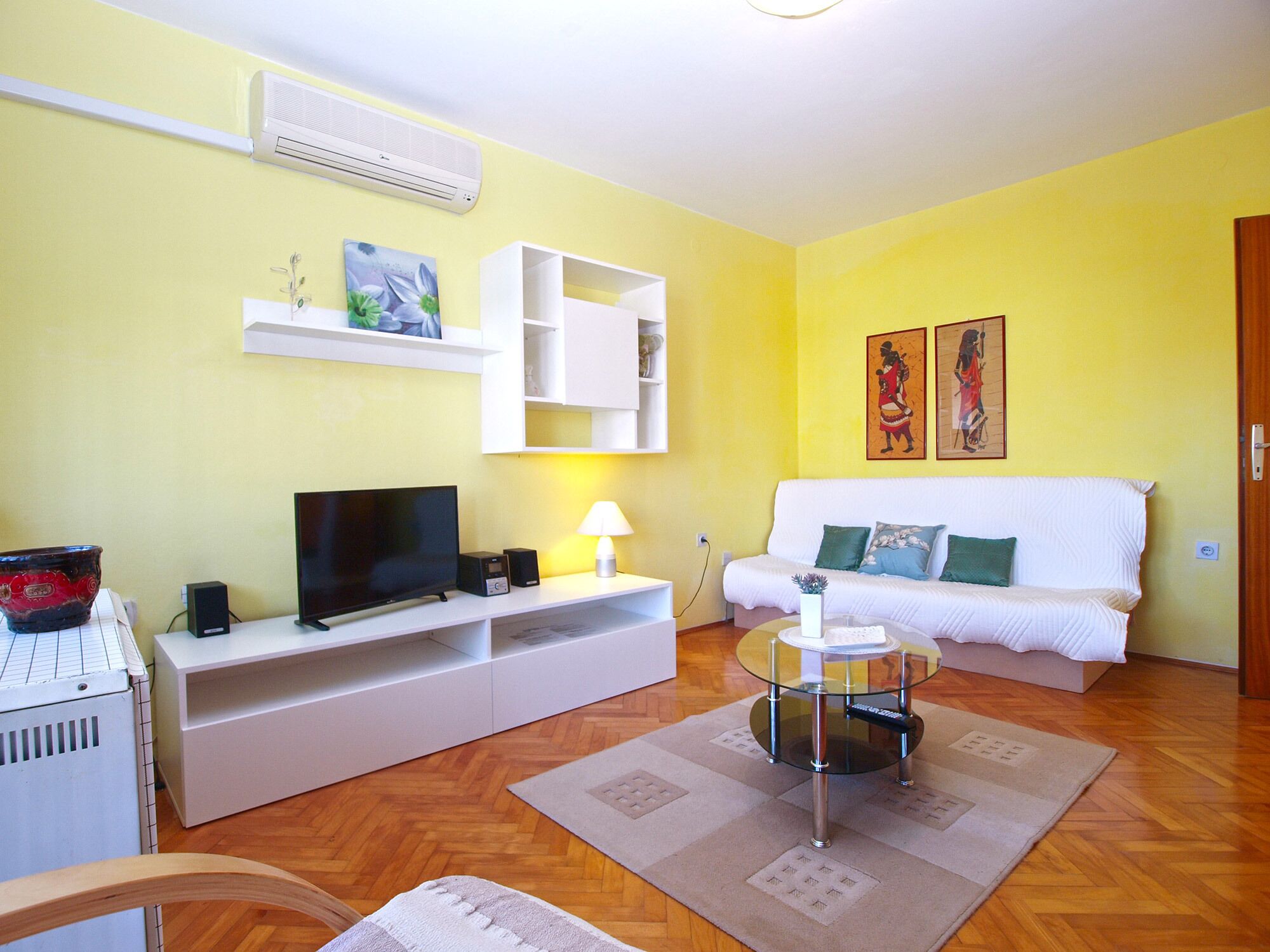 A living room with a white sofa, a glass coffee table, a TV, and a red bowl on a white cabinet.