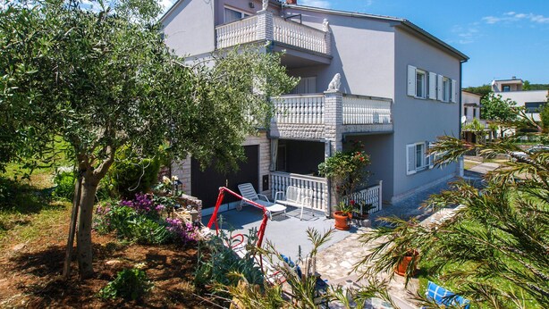 Uma casa de dois andares com varanda, um jardim com diversas plantas e um céu azul límpido.