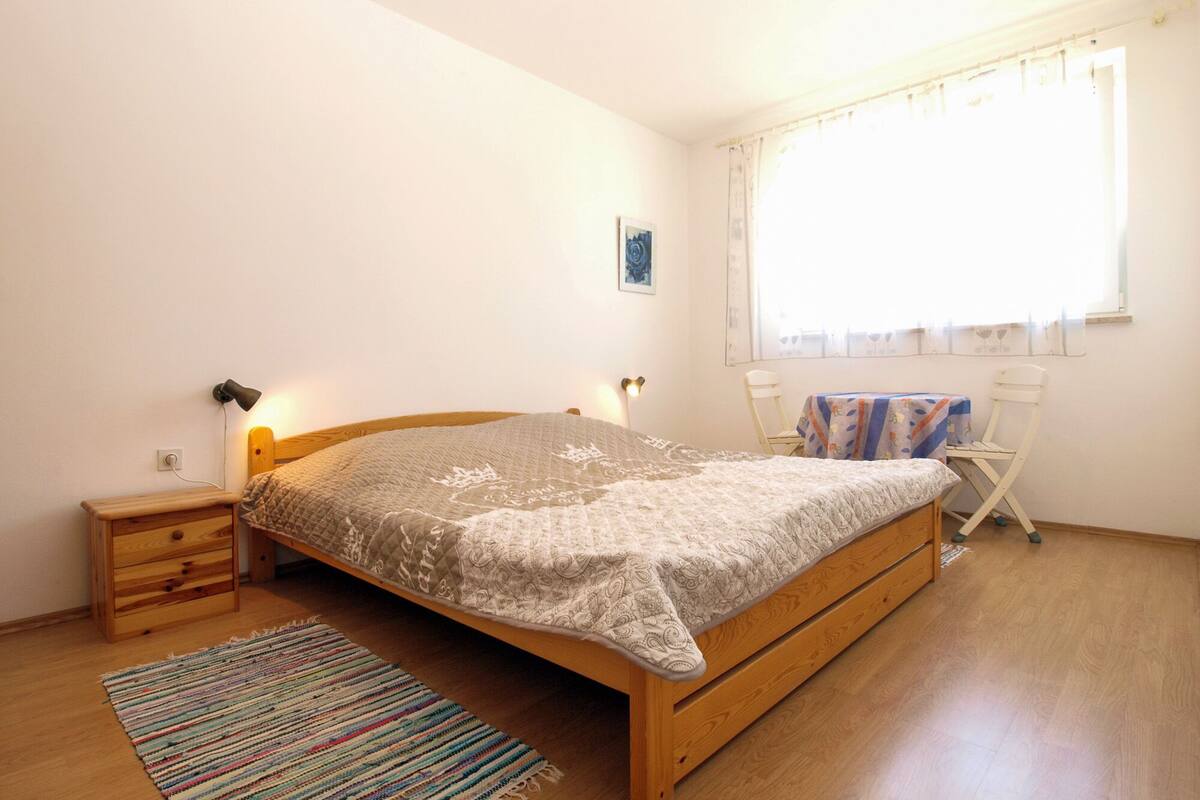 A bedroom with a wooden bed, a bedside table, a small nightstand, a colorful rug, a window with curtains, and a chair.