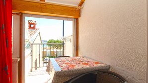 A balcony with a table and chair, a red curtain, and a view of buildings and trees. - Apartment 700 (Rovinj)