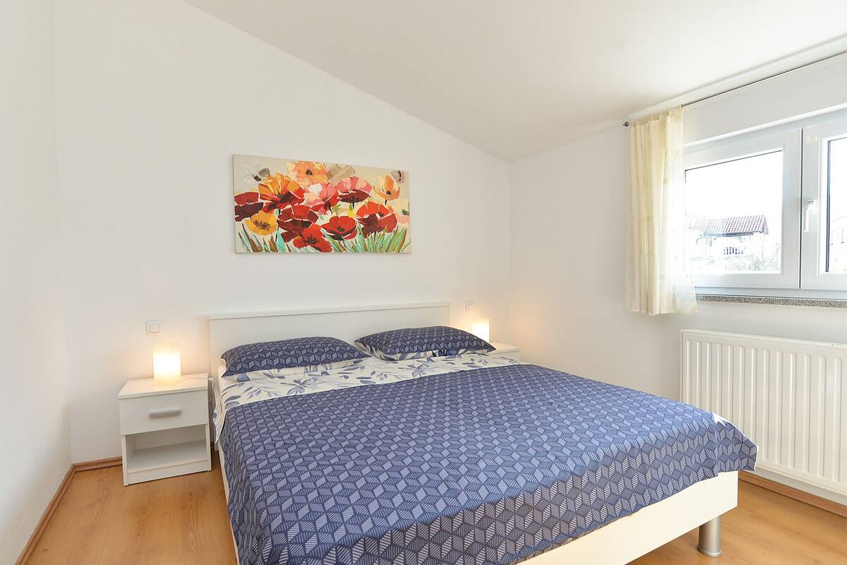 A bedroom with a bed, a nightstand, a window with curtains, and a floral painting on the wall.