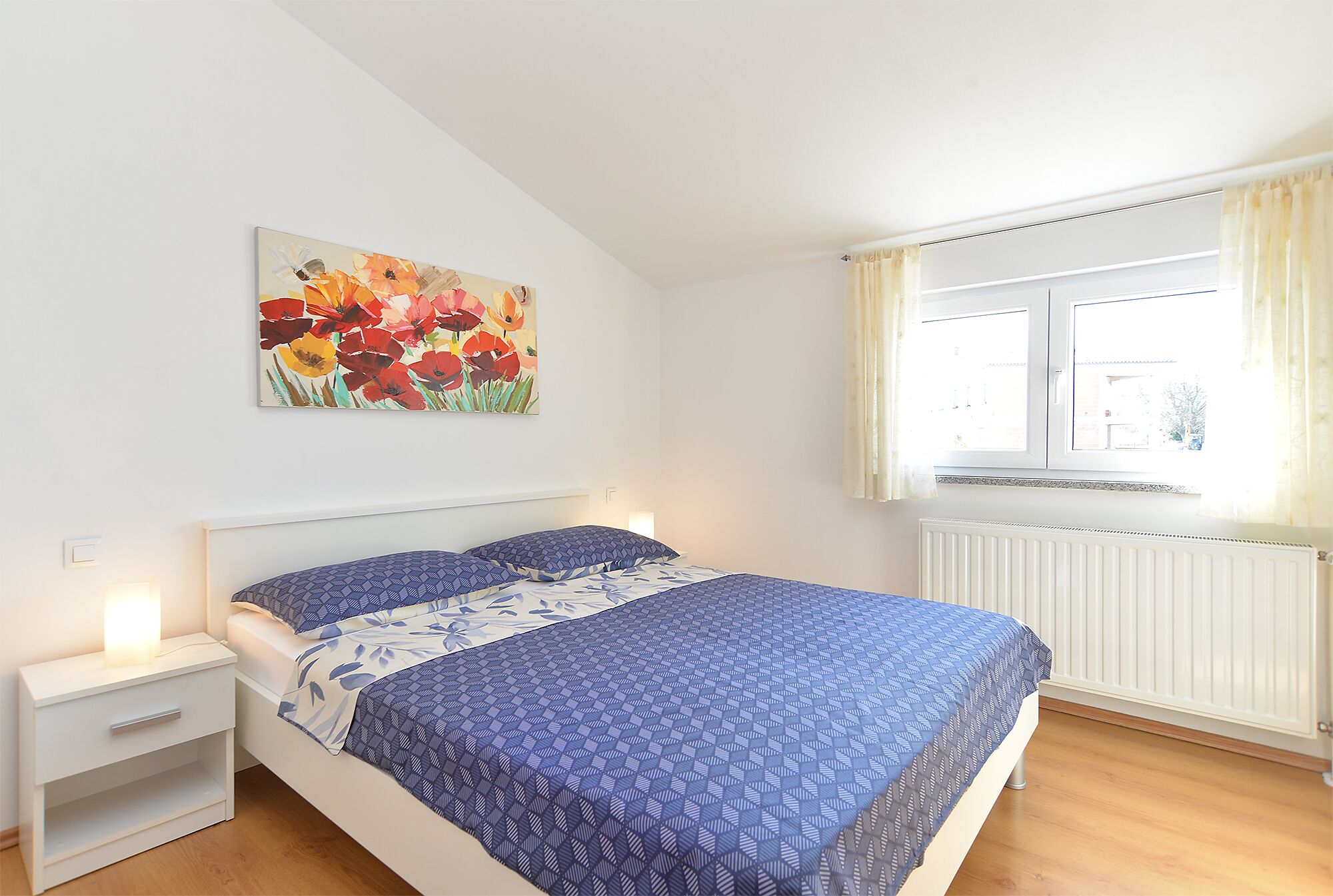A bedroom with a bed, nightstand, and a painting of flowers on the wall.