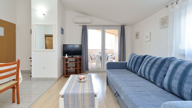 Ein Wohnzimmer mit einer Couch, einem Fernseher auf einem Holzboden, einem Tisch mit gestreiftem Tischläufer und einem Balkon mit Blick.