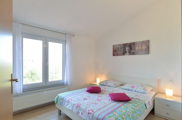 A bedroom with a bed, two bedside tables, a window with curtains, a radiator, and a wall with a decorative piece. - Apartment 719 (Pula)