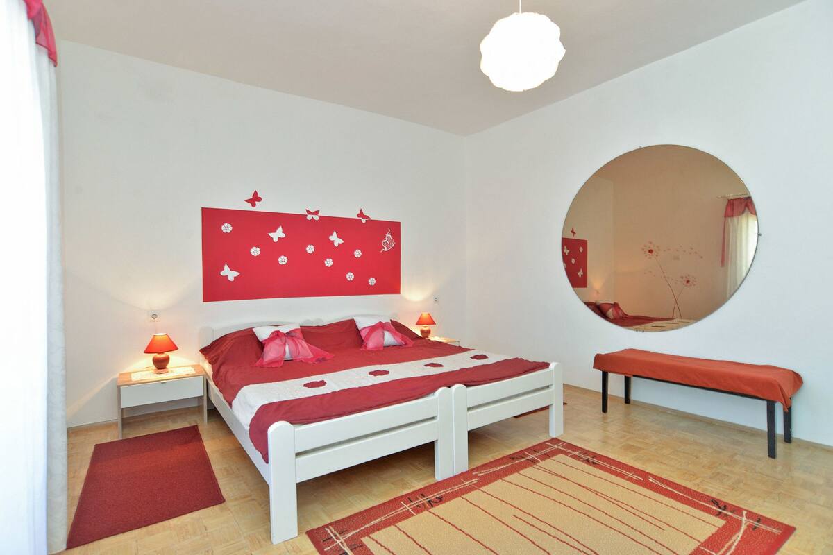 A bedroom with a white bed, red bedding, a round mirror, a red bench, and a wall with butterfly decorations.