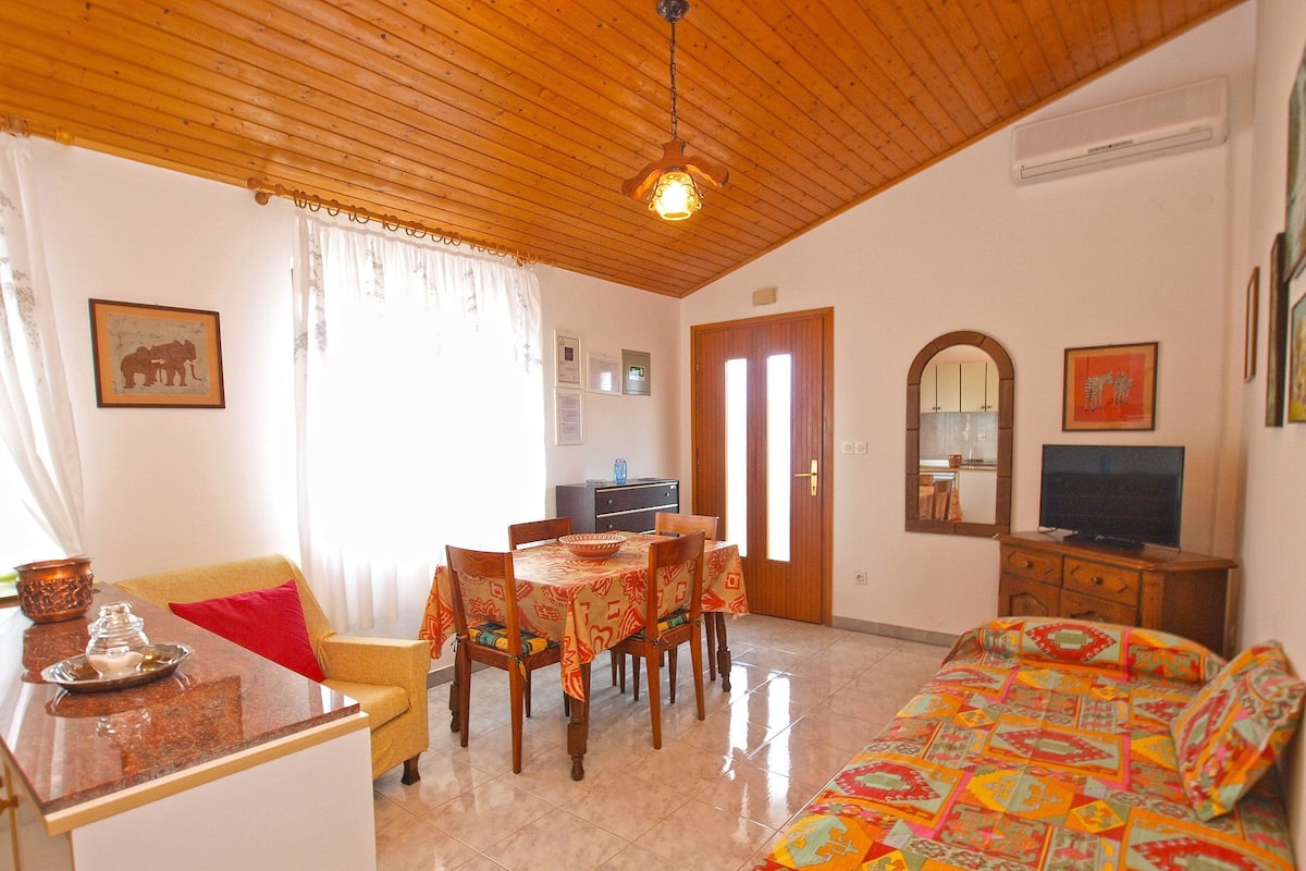 Um quarto acolhedor com teto de madeira, área de jantar com mesa e cadeiras, um sofá, televisão e cozinha visível através de uma porta aberta.