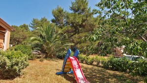 Un parque infantil con tobogán y estructura para escalar, en un área verde con árboles y un edificio al fondo.