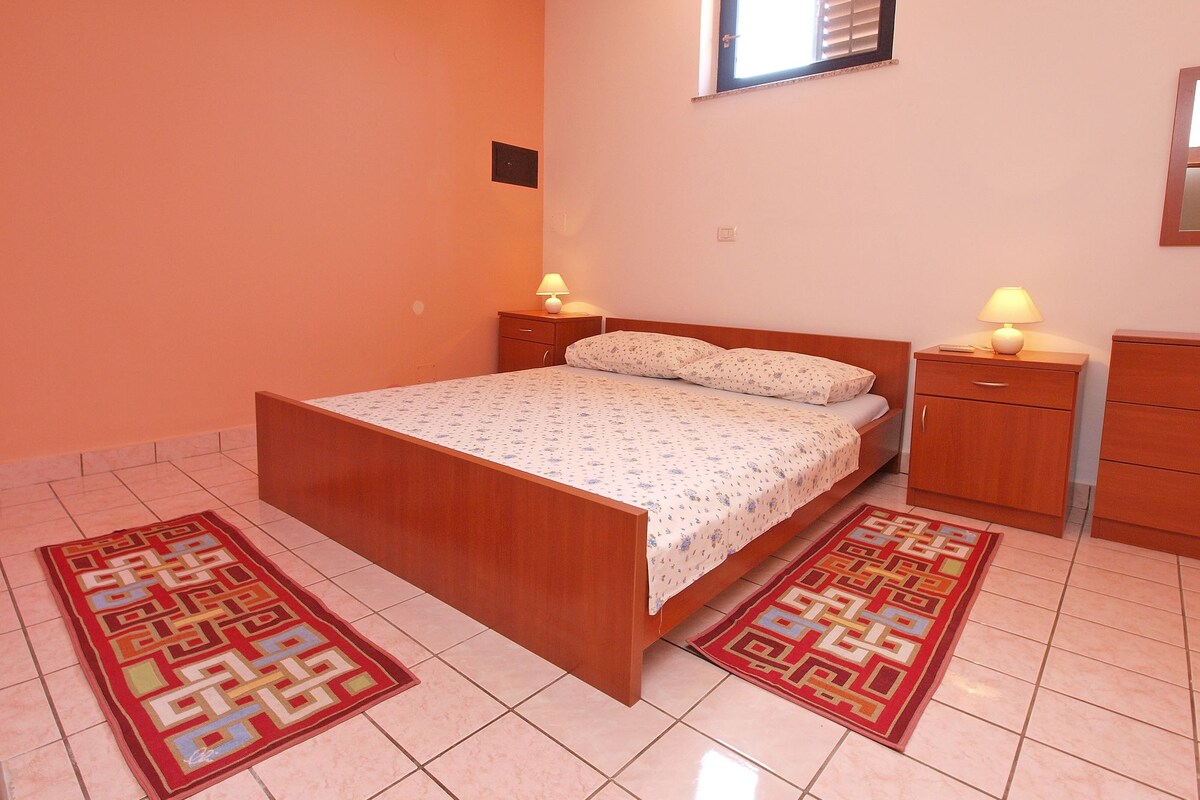 A bedroom with a wooden bed, two bedside tables with lamps, a small window, and a tiled floor.