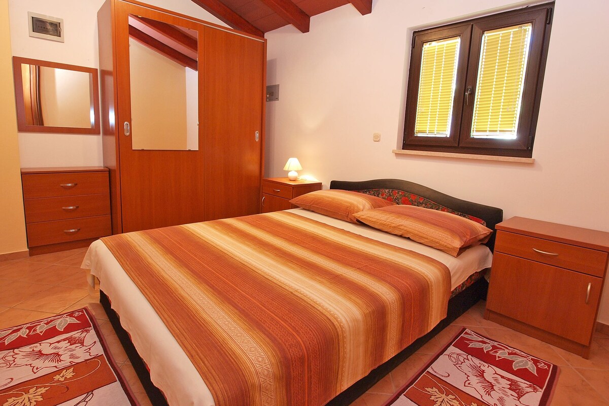 A bedroom with a bed, wooden wardrobe, bedside table, and a window with blinds.