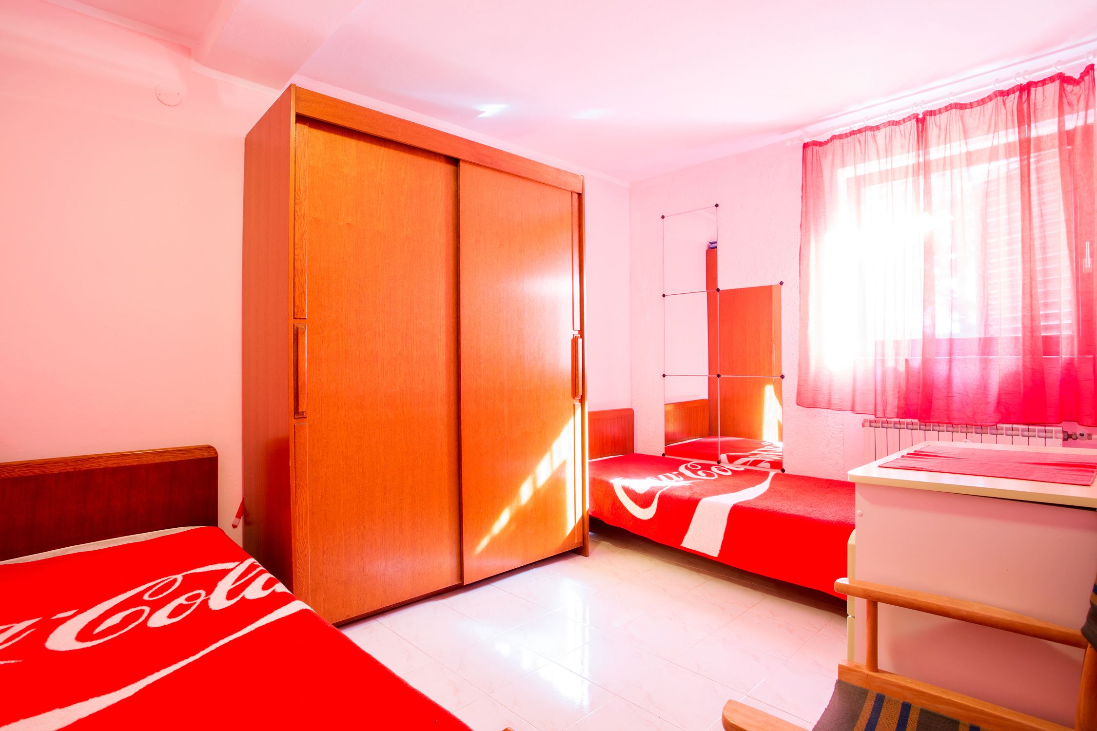 A bedroom with a wardrobe, a bed with a red and white blanket, a desk with a chair, and a window with sheer curtains.
