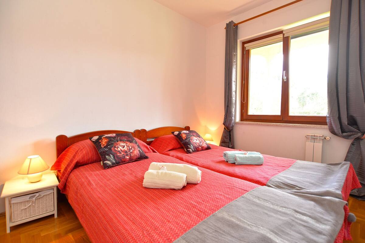Un lit double avec une literie rouge, deux oreillers, une lampe de chevet et une fenêtre avec des rideaux.