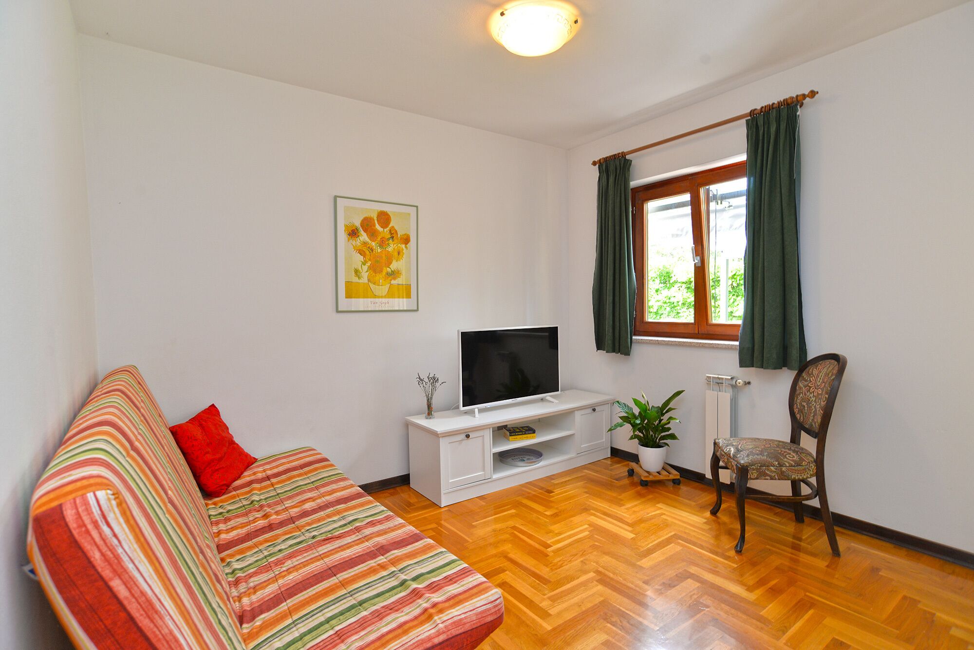 Una sala de estar con un sofá a rayas, un televisor sobre un mueble blanco, una silla, una ventana con cortinas y una planta.