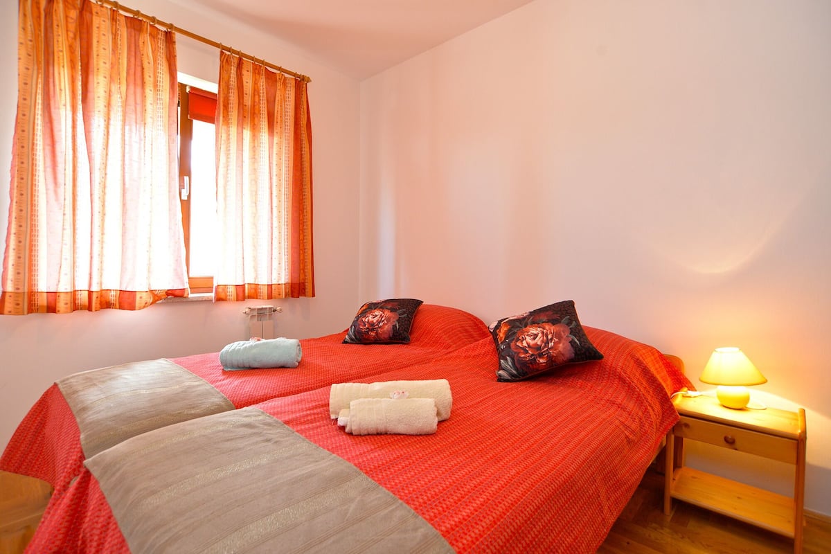 Une chambre à coucher avec une couvre-lit rouge, deux oreillers, une lampe de chevet et une fenêtre avec des rideaux orange.