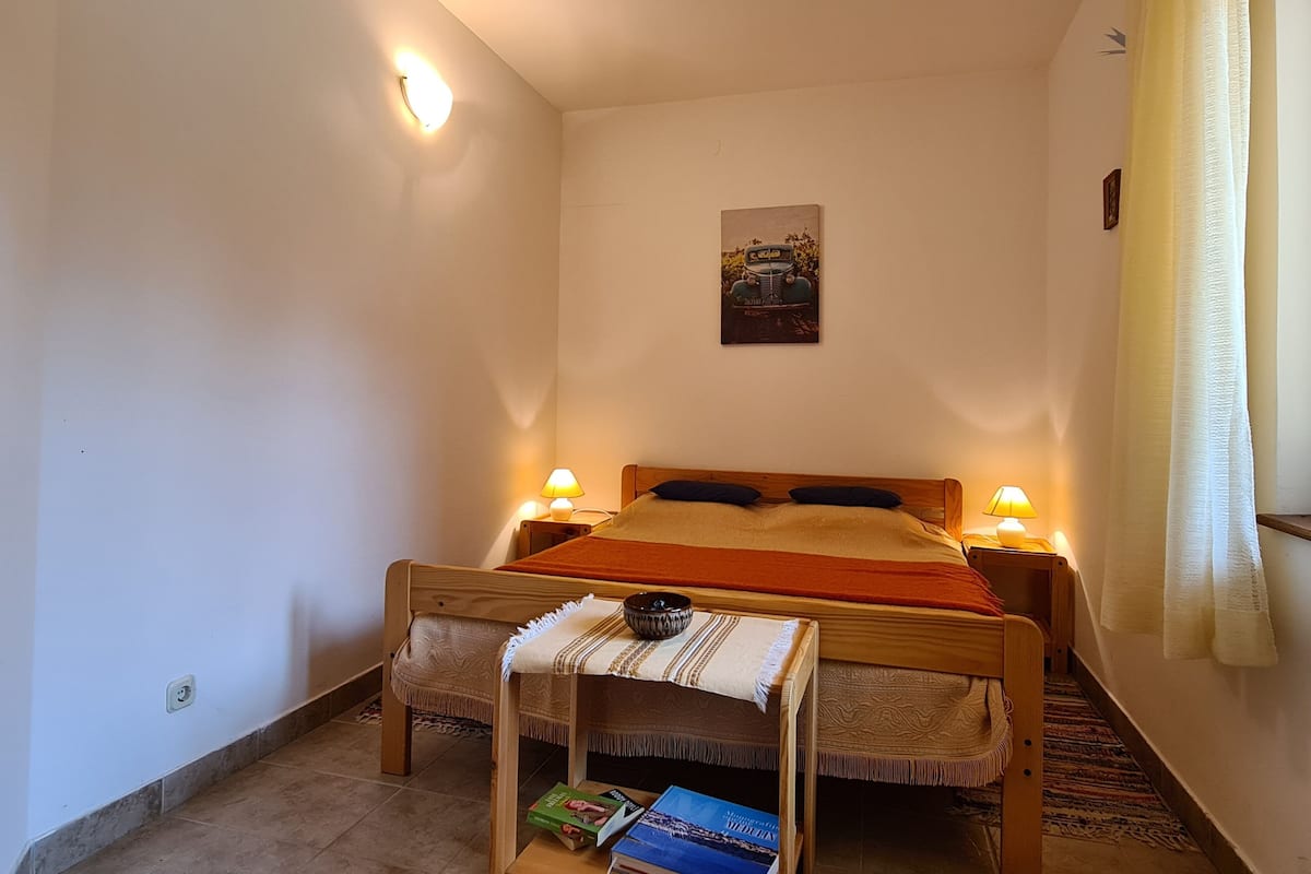 A small, cozy room with a single bed, two bedside lamps, a small table with a bowl and books, and a wall-mounted picture.