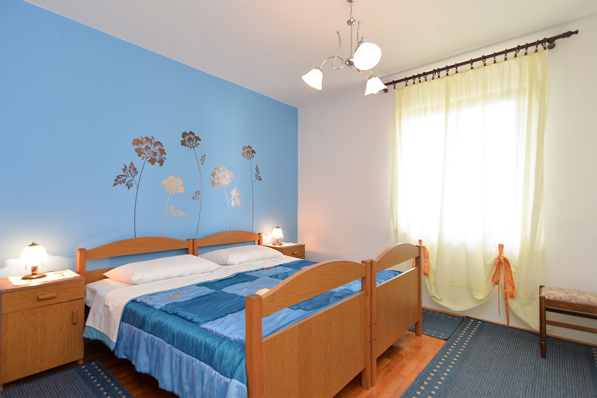 A bedroom with a wooden bed, a nightstand, a lamp, a window with curtains, and a wall with floral decals.